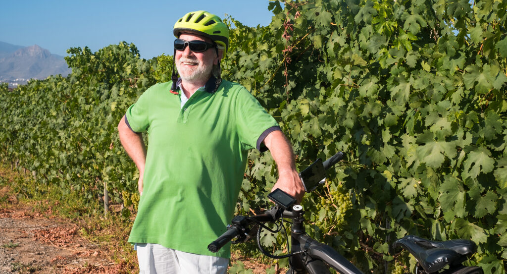 A smiling senior man with white hair and beard standing close to his e bike. Bunches of grapes behind him. Retired elderly caucasian people.Green vineyard in background.